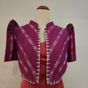 May include: A vibrant purple bolero jacket with white embroidered details and short, puffed sleeves. The jacket features a mandarin collar and a tailored fit. The design includes diagonal white embroidered stripes. The jacket is displayed on a mannequin.