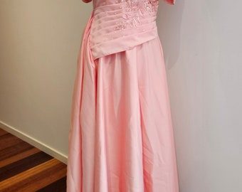 May include: A light pink formal gown with short sleeves and intricate embroidery on the bodice and sleeves. The dress is displayed on a mannequin and features a full skirt. A statement necklace with large, white, pearlescent stones adorns the neckline.