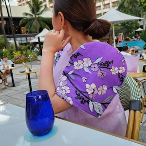May include: A woman wearing a light purple dress with a vibrant purple embroidered shawl featuring floral designs. A blue glass sits on a white table. The background includes palm trees and a hotel.