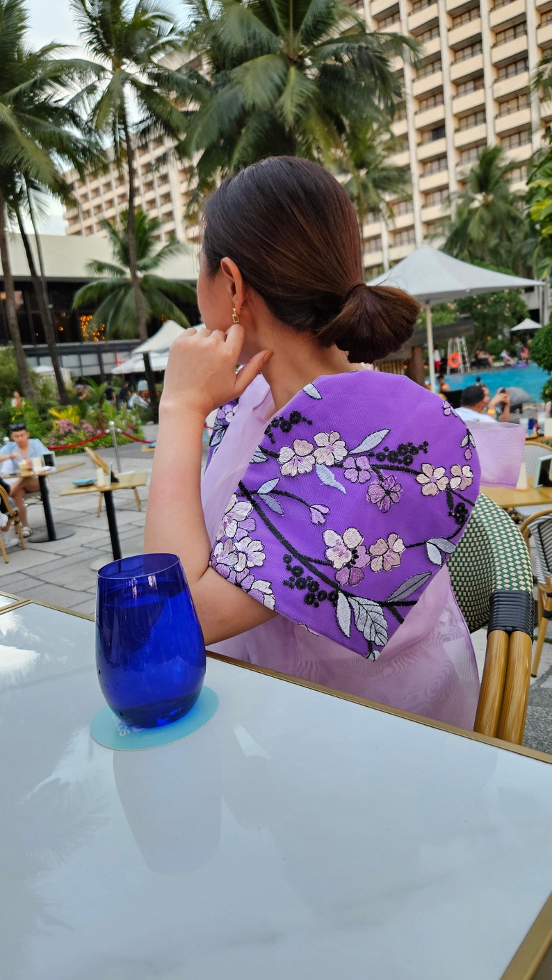 May include: A woman wearing a light purple dress with a vibrant purple embroidered shawl featuring floral designs. A blue glass sits on a white table. The background includes palm trees and a hotel.