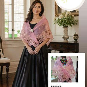May include: A sheer, light pink shawl featuring intricate floral embroidery along the scalloped edges and a central decorative brooch. The shawl is draped over a black sleeveless gown, showcasing a delicate, translucent fabric with detailed stitching.