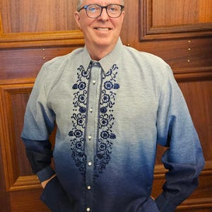 May include: A long-sleeved button-down shirt featuring a blue ombre gradient that transitions from light blue at the shoulders to dark navy at the hem. The front placket is decorated with intricate dark blue floral embroidery patterns.