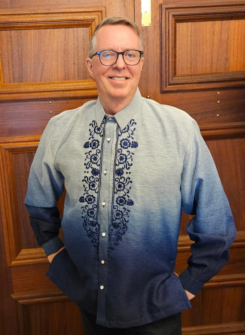 May include: A long-sleeved button-down shirt featuring a blue ombre gradient that transitions from light blue at the shoulders to dark navy at the hem. The front placket is decorated with intricate dark blue floral embroidery patterns.