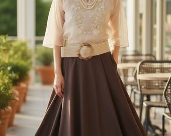 May include: A woman's outfit featuring a cream-colored, embroidered blouse with sheer sleeves, a brown skirt, and a woven belt with a circular buckle. The outfit is accessorized with a pearl necklace.