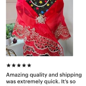 May include: A sheer red shawl featuring intricate white floral embroidery along the scalloped edges. The shawl is fastened at the centre with a decorative brooch made of white pearls and clear crystals, complemented by a matching pearl necklace.