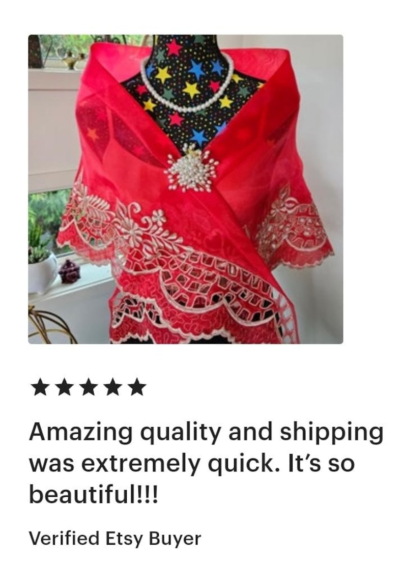 May include: A sheer red shawl featuring intricate white floral embroidery along the scalloped edges. The shawl is fastened at the centre with a decorative brooch made of white pearls and clear crystals, complemented by a matching pearl necklace.