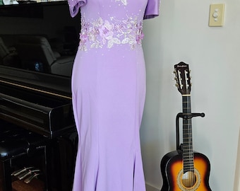 May include: A lilac-colored formal gown with floral and beaded embellishments. The dress features short, puffed sleeves and a mermaid silhouette. A decorative necklace adorns the mannequin. A guitar stands nearby.