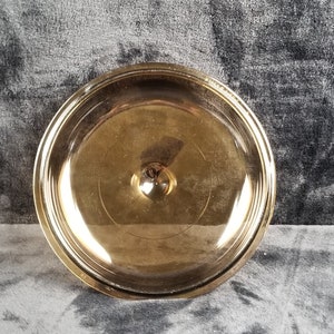 Puede incluir: Una tapa de vidrio transparente con un borde dorado y un pequeño pomo en la parte superior. La tapa es ligeramente abovedada y tiene una superficie lisa y reflectante.