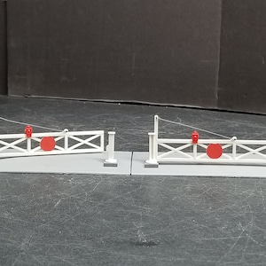 Puede incluir: Dos barreras de paso a nivel de modelo de ferrocarril blanco con señales circulares rojas. Las barreras están cerradas y las señales son visibles.