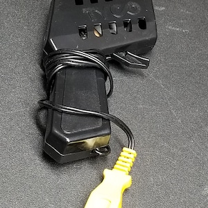 Pode incluir: Controle remoto Tyco preto com um plugue amarelo e um cabo preto. O controle remoto tem um gatilho e um botão no lado.