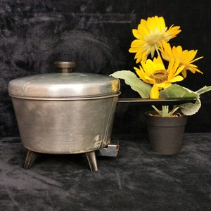 Könnte beinhalten: Eine silberne, dreifüßige elektrische Bratpfanne mit schwarzem Griff und Deckel. Die Bratpfanne steht auf einer schwarzen Oberfläche neben einer Topfpflanze mit gelben Sonnenblumen.