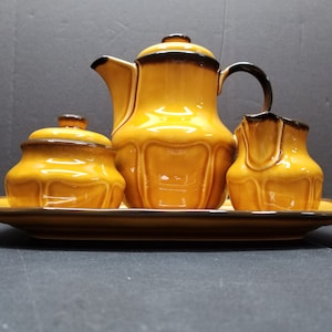 May include: A yellow ceramic tea set with a teapot, creamer, sugar bowl, and tray. The set has a brown trim and a glossy finish.