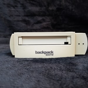 Puede incluir: Un disco duro externo beige con el texto "backpack 800TD" impreso en la parte delantera.