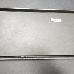 Puede incluir: Bandeja para hornear de metal negro con un revestimiento antiadherente negro extraíble. La bandeja tiene un borde ligeramente curvado y una etiqueta con el número 102.
