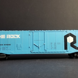 Może przedstawiać: Niebieski model wagonu towarowego z nadrukiem "THE ROCK" i "ROCK 57028" na boku. Wagon ma czarno-białe logo "R" na boku.