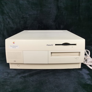 Op de afbeelding: Een beige Power Macintosh 7100/66 computer met een diskettedrive en een stroomkabel. De computer heeft een label met de tekst "Power Macintosh 7100/66" en "PowerPC".