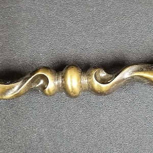 Op de afbeelding: Een messing kastgreep met een decoratief, sierlijk ontwerp. De greep heeft een gedraaide, gebogen vorm met een afgeronde uiteinde aan beide zijden.