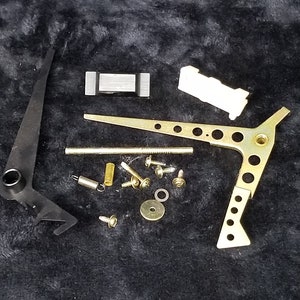 May include: An assortment of clock repair parts, including a black lever, a gold-colored arm with holes, screws, a spring, and a small white plastic piece. The components are arranged on a dark, textured surface.