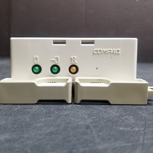 May include: A white plastic COMPAQ computer component with two green LED lights and one orange LED light. The component has two slots with a beige plastic cover.