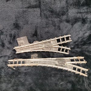 May include: Two silver-colored model train track pieces, one curved and one straight, with a switch mechanism. The pieces are designed to connect to create a track layout.