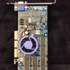 Op de afbeelding: Een blauwe en groene printplaat met een blauwe ventilator en verschillende chips. De printplaat heeft een VGA-connector en een label met de tekst "GIGABYTE".