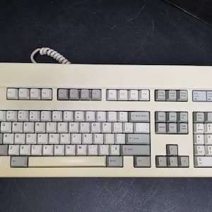 May include: Cream-coloured computer keyboard with grey keys and a coiled cable. The keyboard features a standard QWERTY layout, function keys, a numeric keypad, and indicator lights. Keys are labelled with letters, numbers, and symbols.