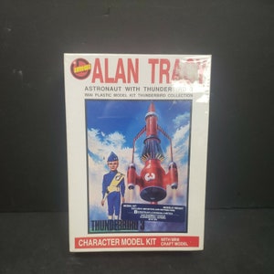 Puede incluir: Una maqueta de plástico Imai en caja con Alan Track, un astronauta, y Thunderbird 3. La caja muestra un cohete rojo y un personaje con uniforme azul sobre un cielo azul. La caja dice "Character Model Kit".