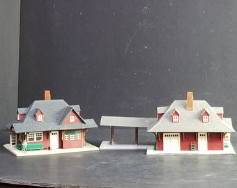 May include: Two model train station buildings with red siding, gray roofs, and white trim. The buildings have a covered platform and a small figure of a person sitting on a bench.