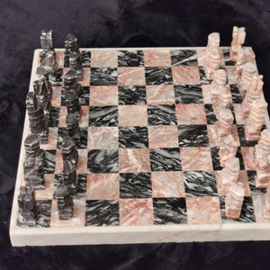 Puede incluir: Un juego de ajedrez de mármol con piezas blancas y negras sobre un tablero de mármol rosa y negro.