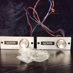 Puede incluir: Dos botones de control de audio de color plateado con el texto "LLOYD'S" y "BRILLIANCE - MIN - MAX" impreso en ellos. Los botones están conectados por cables.