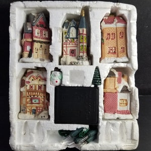 May include: A set of five ceramic Christmas village houses with a snowman and a small tree. The houses are decorated with red, green, and white paint and have lights. The set comes with a battery pack and a string of lights.