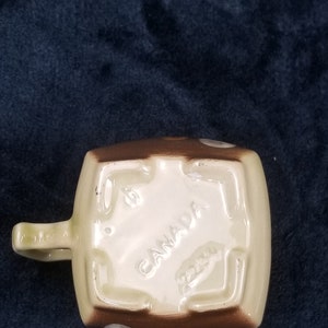 May include: A brown and white ceramic mug with a square shape and a handle. The mug has a brown base with white polka dots and a white top with the word "CANADA" printed in brown.
