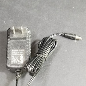 Pode incluir: Adaptador CA preto com um cabo de alimentação preto e um plugue preto. O adaptador tem texto que diz "Entrada: 120V 60Hz 150mA Saída: 12V 1000mA".