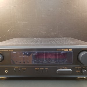 Könnte beinhalten: Ein schwarzer Denon AVR-1404 5.1-Kanal-Surround-Receiver mit digitalem Display und mehreren Knöpfen und Drehreglern.