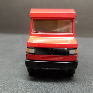 May include: A red toy delivery van with a black front bumper and grill. The van has a rectangular shape with a boxy cabin and a small window. The front of the van features headlights and a Mercedes-Benz logo.