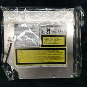 Pode incluir: Uma unidade de CD-ROM Toshiba prateada em um saco de plástico transparente. A unidade tem um rótulo amarelo com o texto "CLASS 1 LASER PRODUCT" e "MADE IN PHILIPPINES".