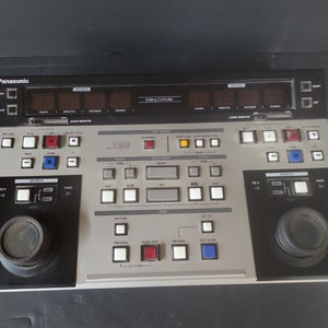 Könnte beinhalten: Ein silberner Panasonic Editing Controller mit einem digitalen Display, Knöpfen und Drehknöpfen. Das Display zeigt "Editing Controller" und einen Zeitcode an. Der Controller hat einen großen Drehknopf links und rechts.