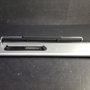 Könnte beinhalten: Eine silberne Laptop-Dockingstation mit einem schwarzen Metallarm und einer Reihe von Anschlüssen an der Vorderseite. Die Dockingstation trägt den Schriftzug "Kensington" an der Seite.