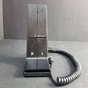 Könnte beinhalten: Schwarze Motorola-Ladestation mit einem Spiralkabel. Die Basis hat einen Ein- und Ausschalter sowie einen Lautstärkeregler. Die Basis ist mit "Motorola" beschriftet und hat ein "AA"-Batteriesymbol.