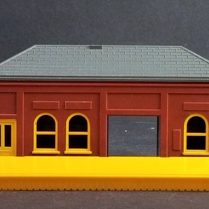 May include: A miniature model of a brick building with a gray roof, yellow trim, and a yellow platform. The building has arched windows and a large door. The model is likely for a train set or diorama.