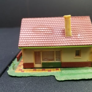 May include: A small, plastic model of a house with a red roof, white walls, and green trim. The house has a chimney and a small yard with a green lawn and a brown brick patio.