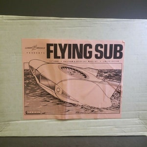 Pode incluir: Uma caixa de cartão branca com uma ilustração rosa e preta de um submarino voador. A caixa diz "Lunar Modules Presents Flying Sub 17" long - Vacuum & Resin Cast Model Kit - Limited Edition".