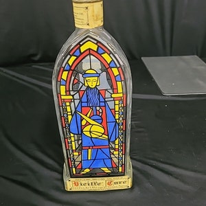 May include: A clear glass bottle with a stained glass design featuring a figure in a blue robe holding a yellow orb. The bottle is labelled "Vicenta Cure" and has a cork stopper.
