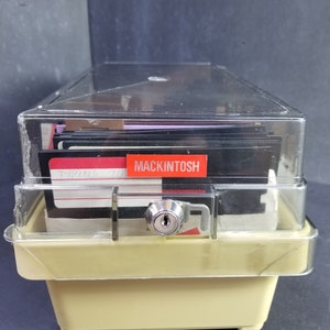 Op de afbeelding: Een beige plastic opbergdoos met een doorzichtig deksel, met daarin een stapel floppy disks. De doos heeft een slot en een sleutel. Een rood label op de bovenste diskette staat "Macintosh".