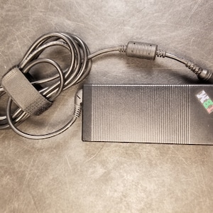 May include: Black laptop power adapter with a ribbed surface and the IBM logo in the corner. The power cord is coiled and secured with a black velcro strap. The adapter is on a dark surface.