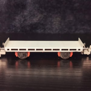 May include: A white model train flatbed car with red wheels and silver accents. The car has a flatbed with a series of slots along the sides.