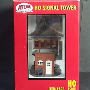 May include: A red box with a white and black logo that says "Atlas Model Railroad Co., Inc." and "HO Signal Tower". The box contains a model of a signal tower with a brown roof, green walls, and a brick base. The tower has two windows and a door. The box also says "HO Scale" and "Item #604".