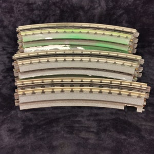 May include: Four curved pieces of model train track stacked on top of each other. The track is made of plastic and has a gray, brown, and green color scheme.