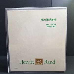 Op de afbeelding: Een beige MS-DOS-handleiding van Hewitt Rand, in een doorzichtige plastic hoes. De omslag toont de bedrijfsnaam en het logo in groen, met de tekst "MS™-DOS MANUAL" ook in groen. Het nummer "585-527F" staat linksonder.