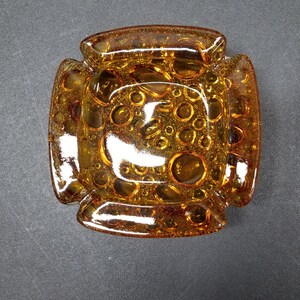 Peut inclure: Cendrier carré en verre ambré avec une surface texturée. Le cendrier a un bord surélevé et un motif de petits cercles.